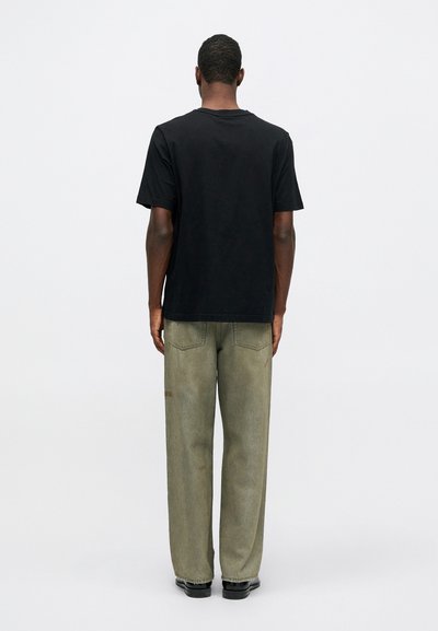 Hombre de pie de espaldas, vistiendo una camiseta negra de manga corta sencilla, vaqueros verdes deslavados y zapatos negros, contra un fondo liso.