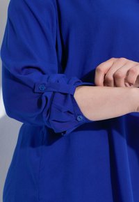 Blusa blu con maniche arrotolate e dettagli con bottoni. Il tessuto appare liscio, con un colore solido e una vestibilità comoda, che sottolinea il comfort.