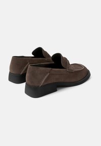 Brown suede loafers with a low black heel, featuring a stitched outline, a rounded toe, and cut-out detail at the heel.