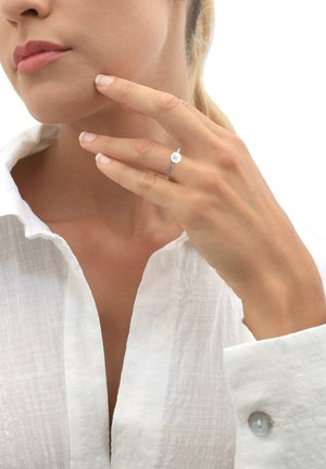 Minimalistinis sidabro žiedas su apvaliu veidu, dėvimas ant rankos, švelniai liečiančios smakrą, derinamas su baltu lino marškinėliu. Sklandi tekstūra.