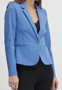 Modrý blazer vyrobený z hladké látky, se zářezovým límcem, zapínáním na jeden knoflík a předními kapsami. Kombinován s černým topem a kalhotami.