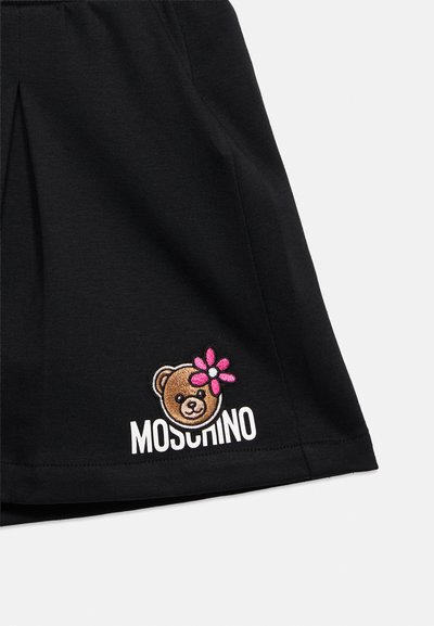 Tissu noir avec une tête d'ours en peluche brodée en marron portant une fleur rose, au-dessus du texte blanc « Moschino » près de l'ourlet inférieur.