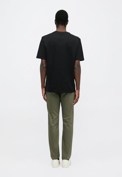 Bărbat stând cu spatele, îmbrăcat cu un tricou negru cu mâneci scurte, pantaloni verde măslin și adidași albi, pe un fundal alb simplu.