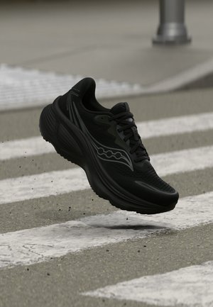 Chaussure de course noire avec logo gris en l’air au-dessus d’un passage piéton avec des bandes blanches et un trottoir gris.