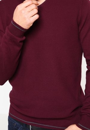 Mężczyzna poprawiający kołnierz bordowego swetra o fakturze z ciemnoszarym wykończeniem, ubrany w ciemnoniebieskie dżinsy i czarny pasek.