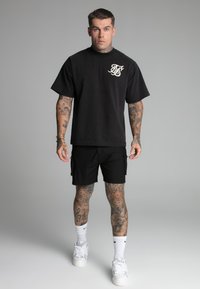 Crna majica kratkih rukava sa detaljem loga, u kombinaciji s crnim cargo šortsevima i bijelim sportskim cipelama, na modelu s tetovažama.