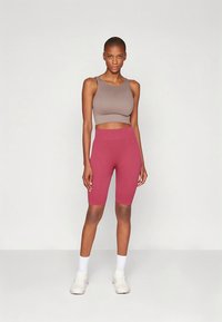 Sporta crop top brūnā krāsā un augstas jostasvietas rozā riteņbraukšanas šorti, abi izgatavoti no elastīgas auduma, kopā ar baltiem sporta apaviem un zeķēm.