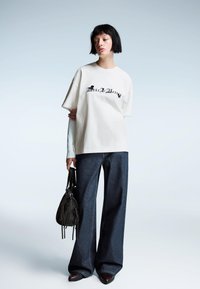 Overmaatse beige katoenen t-shirt met zwarte tekst, gecombineerd met wijde donkerblauwe jeans en een kleine zwarte handtas.