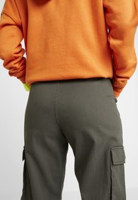 Gröna cargobyxor med sidofickor, i kombination med en orange hoodie. Tyget verkar mjukt med en avslappnad passform som framhäver sömnadsdetaljer.