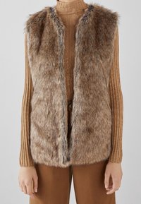 Vesta z umělé kožešiny v hnědých tónech s měkkým, texturovaným povrchem. Má límec, otevřený předek a je zkombinována s hnědým žebrovaným svetrem.