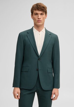 Junger Mann trägt einen dunkelgrünen maßgeschneiderten Anzug und ein weißes Strickshirt mit Reißverschluss, steht vor einem schlichten hellen Hintergrund.