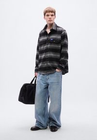 Μαύρο και γκρι ριγέ πουκάμισο φανέλας με τσέπες με κουμπιά, συνδυασμένο με ανοιχτό μπλε τζιν παντελόνι και μαύρα παπούτσια χωρίς κορδόνια, κρατώντας μια μαύρη τσάντα.