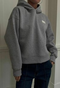 Felpa grigia con cappuccio realizzata in materiale morbido, dotata di una tasca frontale e polsini a coste. Presenta un piccolo logo rettangolare sul petto.