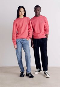 Két modell azonos korall színű pulóverben, apró logókkal. Az egyik farmernadrágot visel, a másik fekete nadrágot. Mindketten sportcipőt hordanak.