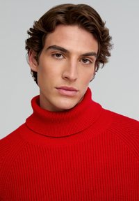 Joven con cabello castaño ondulado, vistiendo un suéter de cuello alto rojo, mirando hacia adelante con una expresión neutral sobre un fondo gris claro.