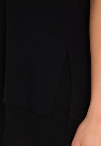 Svart tyg topp med en slits på sidan, mjuk textur och något lös passform. Nederkanten är rak utan dekorativa inslag.