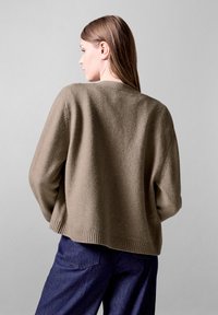 Brauner strukturierter Pullover mit angeschnittenen Schultern und geripptem Saum, kombiniert mit dunklen Jeans. Die Ansicht ist von hinten.