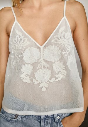 Camisole en maille blanche avec décolleté en V, ornée de broderies florales complexes. Bretelles spaghetti et coupe ample, associée à un short en denim bleu clair.