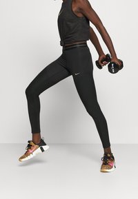 Czarny sportowy tank top z złotym logo, wysokowarstwowe czarne legginsy z teksturowanymi akcentami oraz kolorowe buty sportowe z wzorem.