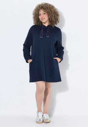 Frau mit lockigem Haar, die ein marineblaues Hoodie-Kleid mit langen Ärmeln und silberne Sneakers trägt, steht mit den Händen in den Taschen vor einer weißen Wand.