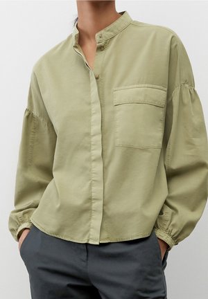 Osoba ubrana w luźną, jasnooliwkową koszulę na guziki z kieszenią na piersi i bufkowymi rękawami, w połączeniu z ciemnoszarymi spodniami.