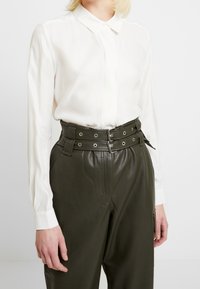 Bílá dlouhá košile s límečkem ve tvaru špičky, v kombinaci s tmavě zelenými kalhotami z umělé kůže s vysokým pasem a dvojitým páskem.