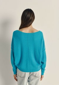 Turkis oversize sweater lavet af blødt stof, med vid halsudskæring, sænkede skuldre og ribbede manchetter, parret med lyse jeans.