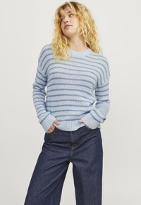 Világoskék csíkos pulóver puha anyagból, hosszú ujjú és bordás mandzsettával, sötét farmernadrággal párosítva. Semleges háttér.