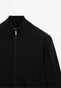 Pull à fermeture éclair en maille noire avec col montant côtelé, fermeture éclair argentée et discrète étiquette de marque à l'intérieur du col.