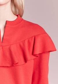 Sweatshirt corail avec un col côtelé, présentant un grand volant sur la poitrine. Le tissu semble doux, avec une texture lisse.