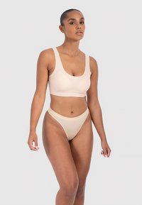 Beige naadloze beha met brede bandjes, diepe halslijn en lichte compressie. Bijpassende hooggetailleerde string met elastische tailleband. Glad oppervlak.