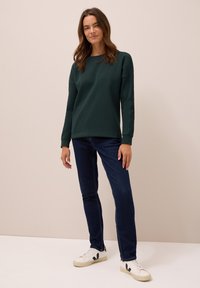 Mörkgrön sweatshirt med avslappnad passform, rund halsringning och långa ärmar, ihop med mörkblå slim fit jeans och vita sneakers.