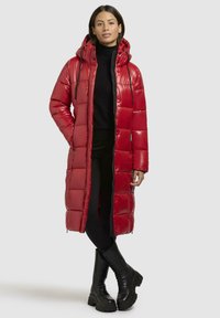 Rød pufjakke med kviltet mønster, hætte og lynlås foran. Bæres med sort turtleneck og strukturerede sorte knæhøje støvler.