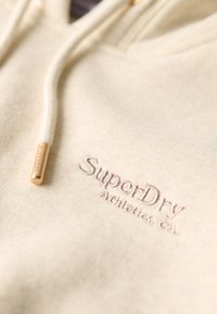 Hoodie crema realizzato in tessuto morbido, con cordino dotato di punta in metallo dorato marchiata. "SuperDry Athletics Co." ricamato in un colore a contrasto.