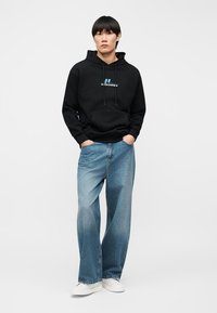 Homme portant un sweat à capuche noir avec un logo, un jean à jambes larges bleu et des baskets blanches, debout les mains dans les poches sur un fond blanc.