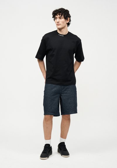 Νέος άνδρας με σγουρά μαλλιά που φοράει μαύρο μπλουζάκι και σκούρο μπλε σορτς με λεπτές ρίγες, στέκεται με τα χέρια πίσω από την πλάτη σε λευκό φόντο.