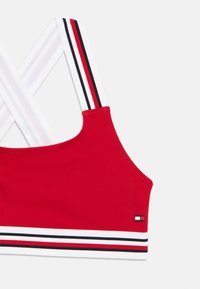 Κόκκινο αθλητικό σουτιέν με σταυρωτούς λευκούς ιμάντες με κό κκινες, μαύρες και λευκές ρίγες, και επωνυμία στο πλάι. Λείο ύφασμα.