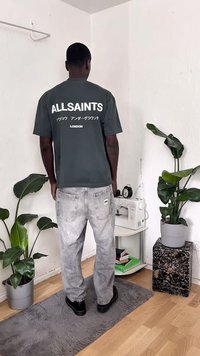 Γκρι βαμβακερό t-shirt με το "ALLSAINTS" σε λευκή γραφή στην πλάτη, συνδυασμένο με ανοιχτό γκρι τζιν και μαύρα παπούτσια. Εσωτερικός χώρος με φυτά.