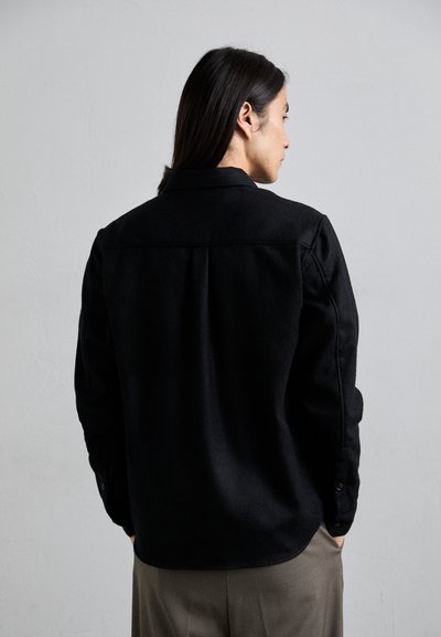 Black wool jacket with a collared neck, button cuffs, and a smooth texture, featuring a slightly curved hem and back seam details.