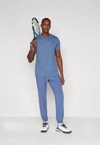 Zils sporta īsās piedurknes krekls ar saskaņotiem sporta biksēm, izgatavots no viegla auduma. Modelis tur tenisa raketi un valkā baltus sporta apavus.