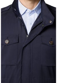Granatowa kurtka z zamkiem, z dwoma kieszeniami na piersiach z guzikami i wysokim zapięciem, noszona na jasnej, wzorzystej koszuli.