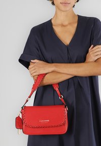 Borsa a tracolla rossa in pelle con dettagli a cucitura intrecciata, tracolla regolabile e hardware dorato. Presenta un piccolo charm attaccato.