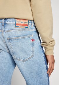 En person med beige ärmar som bär ljusblå Diesel-jeans, och visar bakfickan samt lädermärkeslappar ovanför midjebandet.