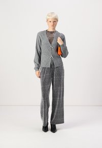 Hallide hallhargiga, millel on kohandatav sulgemine, koos hallide tekstuuriga laienevate pükstega. Mudel hoiab käes väikest oranži kotikest.