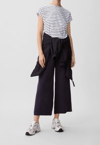 Camiseta de rayas blancas y marinas, pantalones anchos negros, chaqueta negra atada a la cintura y zapatillas grises. Conjunto casual y cómodo.