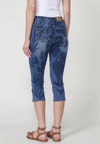 Capris en denim avec un motif floral bleu, cinq poches et fermetures éclair aux chevilles. Le matériau est extensible et ajusté, avec une taille mi-haute.