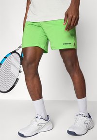 Limegrüne Sportshorts mit Seitenschlitzen, schwarzem "HEAD"-Logo, kombiniert mit weißen Schuhen und weißen Knöchelsocken, während sie einen Tennisschläger halten.