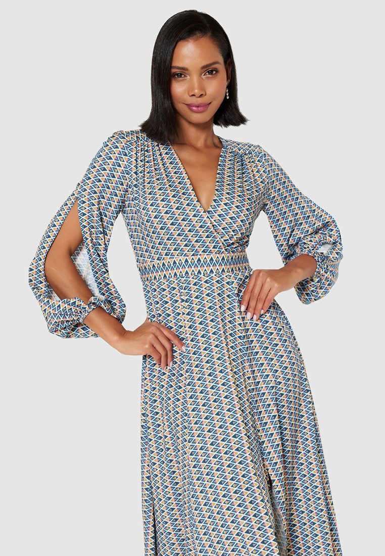 Kobieta w wzorzystej sukience z długim rękawem i rozciętymi rękawami, głębokim dekoltem w kształcie litery V, z zaznaczoną talią oraz geometrycznym wzorem w kolorze niebieskim, żółtym i białym, pozująca z pewnością siebie.