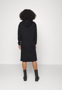 Henkilö, jolla on kihara hius, pukeutuneena mustaan huppari-mekkoon ja mustiin nilkkasaappaisiin, seisoo vaaleaa taustaa vasten.