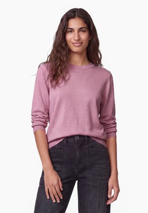Junge Frau mit langen, welligen Haaren, die einen pinkfarbenen Langarmpullover und dunkle, hoch taillierte Hosen trägt, steht vor einem weißen Hintergrund.